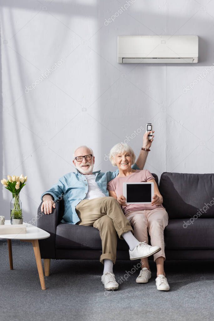Smiling senior couple showing digital tablet and holding remote controller of air conditioner at home