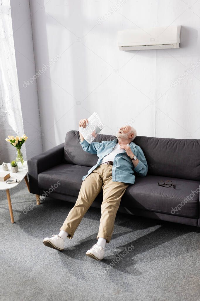 Senior man waving newspaper while suffering from heat at home 