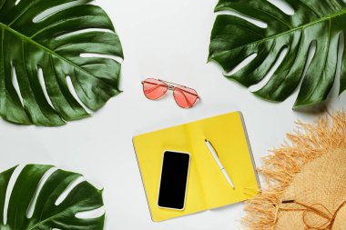 Yeşil palmiye yapraklarının üst görüntüsü, hasır şapka, güneş gözlüğü ve beyaz arkaplanda akıllı telefon olan sarı not defteri.