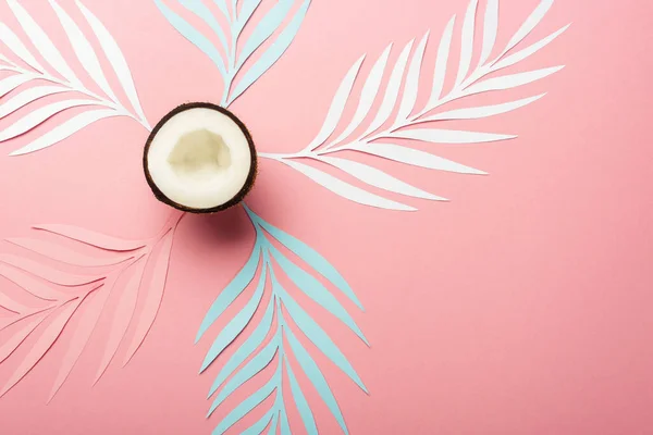 top view of white, pink and blue paper cut palm leaves and coconut half on pink background