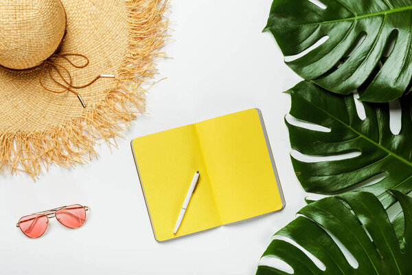 top view of green palm leaves, straw hat, sunglasses and yellow notepad on white background