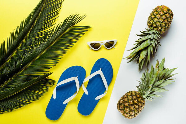 top view of green palm leaves, pineapples, sunglasses and blue flip flops on white and yellow background