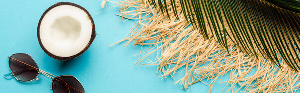 top view of green palm leaf, coconut half, sunglasses and straw hat on blue background, panoramic shot