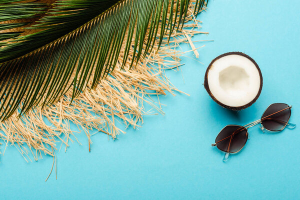 top view of green palm leaf, coconut half, sunglasses and straw hat on blue background