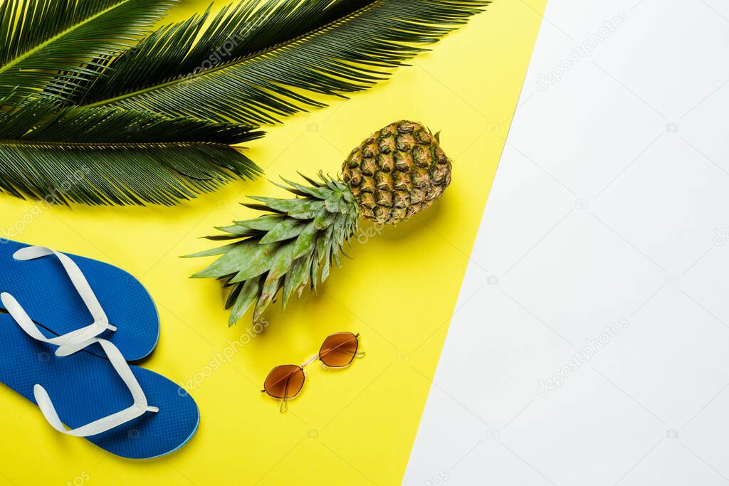 top view of green palm leaves, pineapple, sunglasses and blue flip flops on white and yellow background