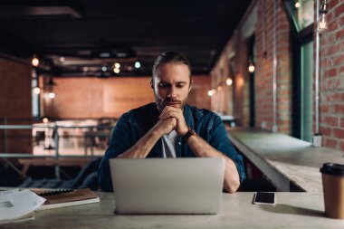 Elleri sımsıkı bağlı işadamı, kağıt bardağın yanındaki dizüstü bilgisayara bakıyor ve masasında boş ekran olan akıllı telefona bakıyor.