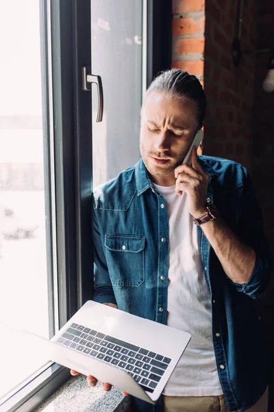 ノートパソコンを持っていて窓の近くのスマートフォンで話しているハンサムなビジネスマン — ストック写真