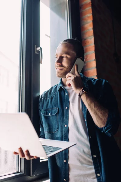 ノートパソコンを持ち窓の近くのスマートフォンで話しているビジネスマンの — ストック写真