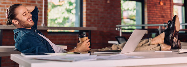 panoramic crop of cheerful businessman holding paper cup and looking at laptop in office
