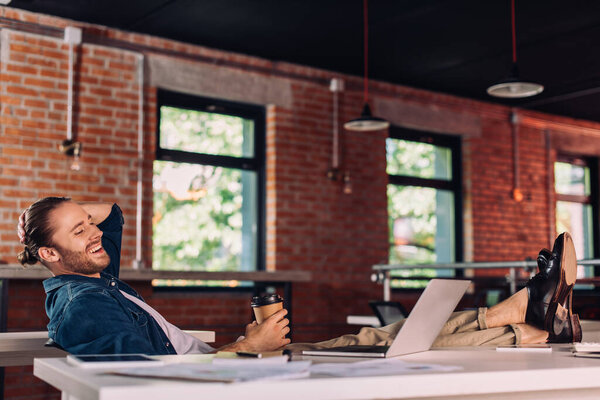 selective focus of cheerful businessman holding paper cup and looking at laptop in office