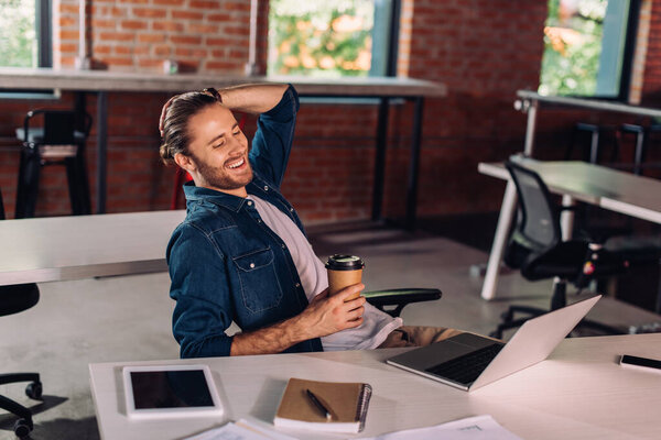 happy businessman holding paper cup and using laptop near smartphone and digital tablet with blank screen on desk