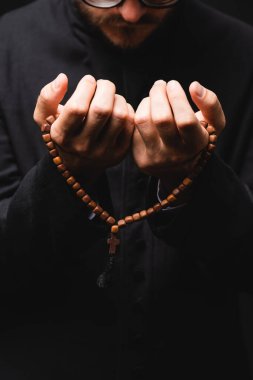 cropped view of pastor holding rosary beads in hands and praying isolated on black  clipart