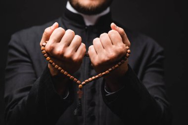 selective focus of pastor holding rosary beads in hands and praying isolated on black  clipart