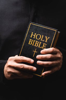 partial view of priest holding holy bible in hands isolated on black  clipart