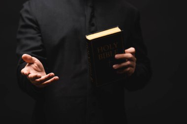 partial view of priest holding holy bible while gesturing isolated on black  clipart