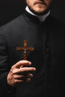 selective focus of priest holding wooden cross isolated on black  clipart