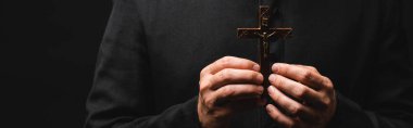 panoramic crop of pastor holding wooden cross isolated on black  clipart