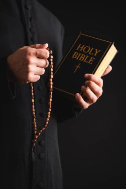 cropped view of priest holding holy bible and rosary beads in hands isolated on black  clipart