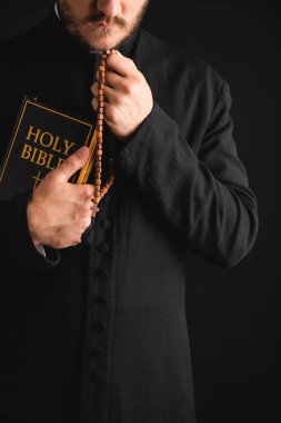 cropped view of bearded priest holding holy bible and rosary beads in hands isolated on black  clipart