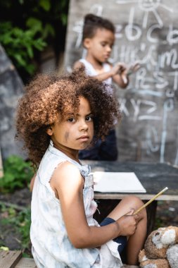 Çocuk ve karatahtanın yanında otururken elinde kalem tutan Afrikalı Amerikalı çocuğun seçici odak noktası 