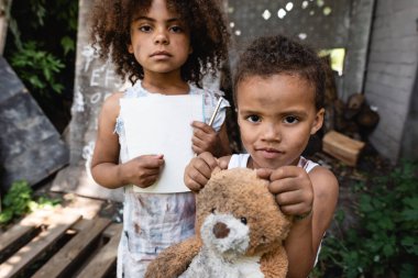 Zavallı Afrikalı Amerikalı çocuğun elinde boş kağıt ve kalemle kardeşinin yanında oyuncak ayıyla durduğu seçici odak noktası. 