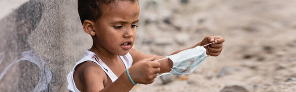 panoramic shot of poor african american kid holding dirty medical mask outside
