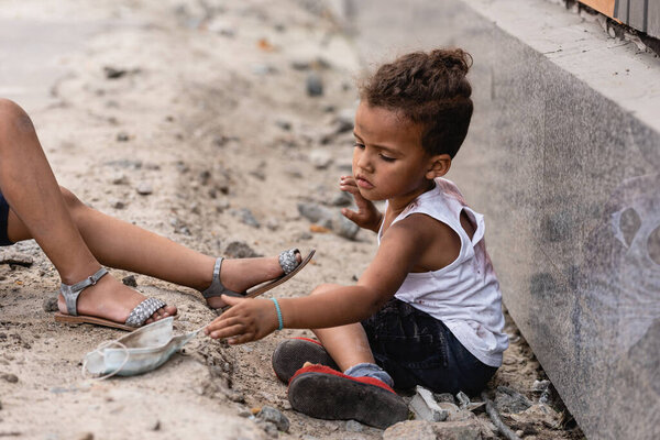 poor african american boy reaching dirty medical mask near sister on ground