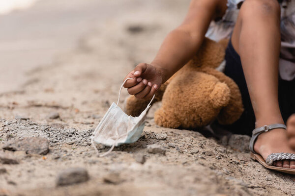 cropped view of poor african american child touching dirty medical mask near teddy bear on ground
