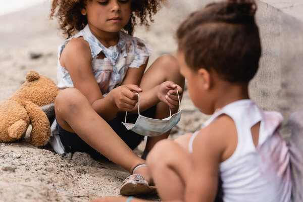 selective focus of curly african american child holding dirty medical mask near poor brother sitting on ground