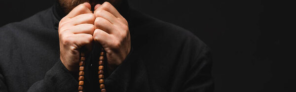 panoramic crop of priest holding rosary beads in hands isolated on black 