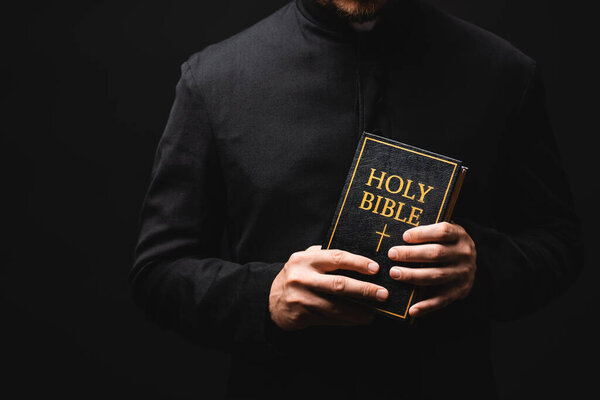 cropped view of priest holding holy bible in hands isolated on black 