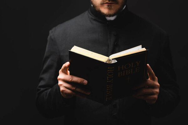 partial view of bearded pastor reading holy bible isolated on black 