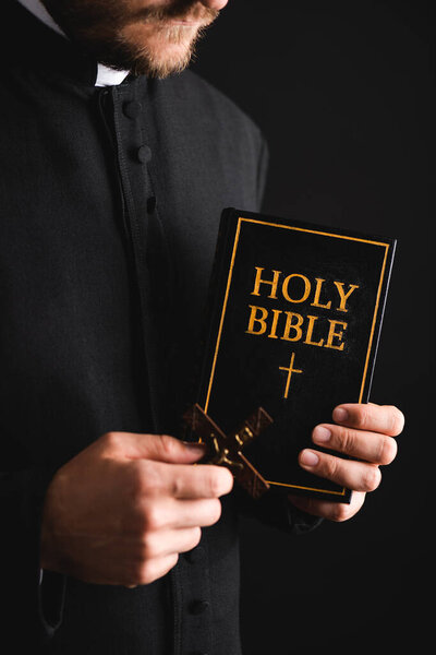 cropped view of bearded priest holding cross and holy bible isolated on black 