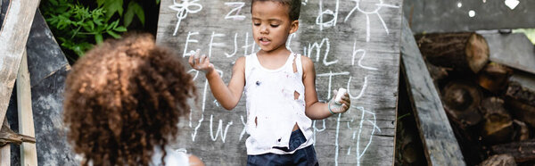 panoramic shot of poor african american kid standing near chalkboard and curly sister 