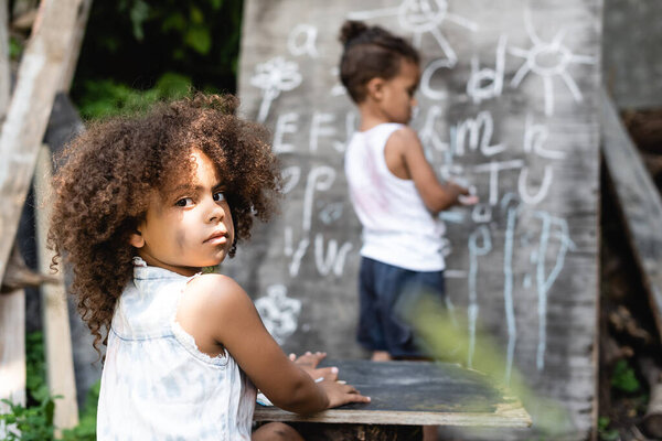 selective focus of curly african american kid sitting near table, boy and chalkboard 