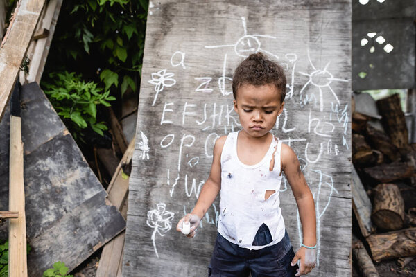 sad african american boy in torn clothes holding chalk near chalkboard 