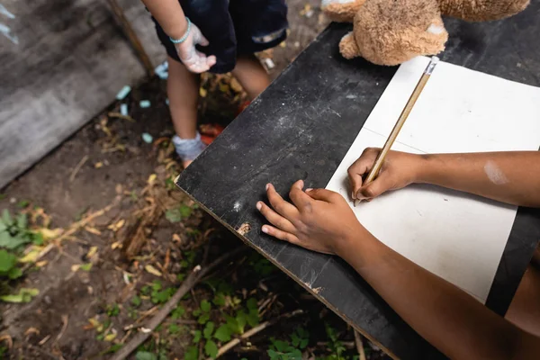 Częściowy Widok Afrykańskiego Dziecka Trzymającego Ołówek Podczas Pisania Pobliżu Dziecka — Zdjęcie stockowe