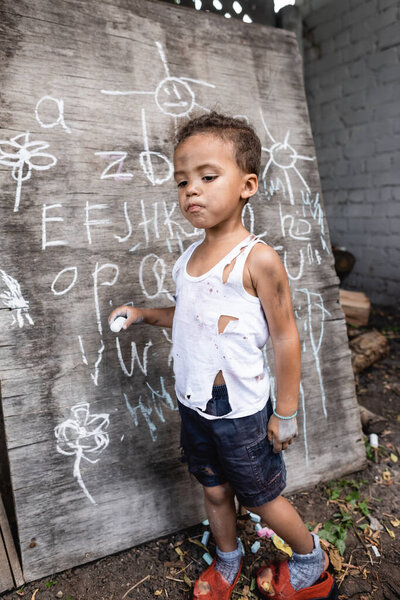 African american child in torn clothes holding chalk near chalkboard 