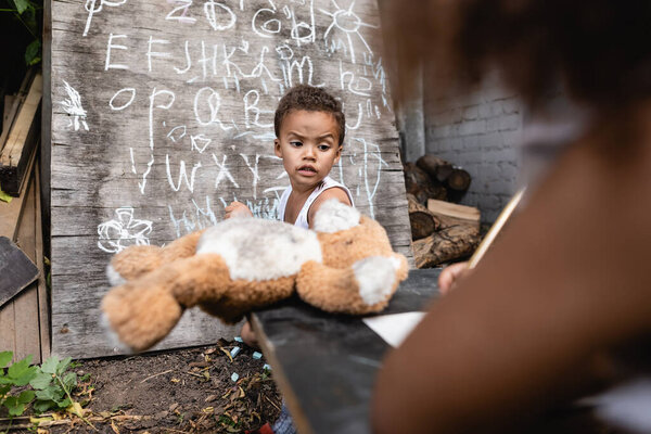 selective focus of african american boy holding soft toy near kid and chalkboard 