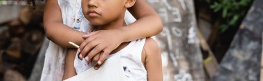 panoramic crop of african american kid holding pencil and hugging poor brother in torn clothes  clipart