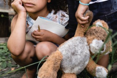 cropped view of poor african american kid holding notebook near child standing with dirty soft toy clipart