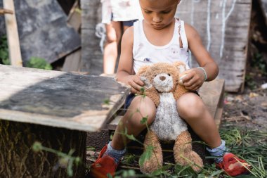 Yırtık elbiseli, yumuşak oyuncakla oturan zavallı Afrikalı Amerikalı çocuğun seçici odağı 