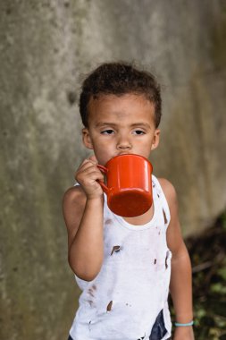 Dirty african american boy holding metal cup in slum  clipart