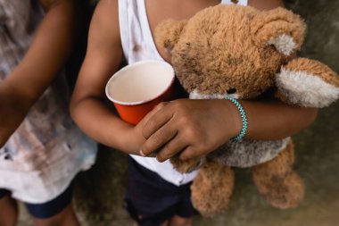 Cropped view of dirty african american children with teddy bear and cup begging alms on urban street  clipart