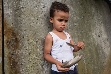 Üzgün, yoksul, Afro-Amerikan çocuğu elinde metal tabak ve kaşık tutarken sokak ortasında dilenirken. 