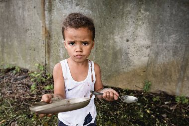 Dissatisfied african american boy holding dirty plate and spoon and looking at camera on urban street  clipart