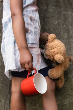 Cropped view of poor african american child holding teddy bear and metal cup on urban street  clipart