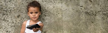 Panoramic shot of african american child holding purse near concrete wall on urban street  clipart
