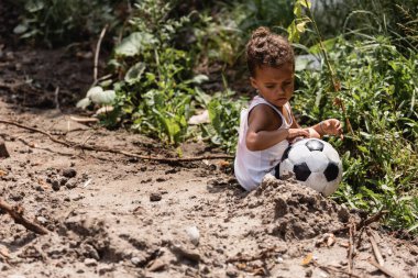 Selective focus of poor african american boy sitting near soccer ball and plants on dirty road  clipart
