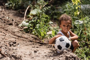 Zavallı Afrikalı Amerikalı çocuk kirli yoldaki bitkilerin yanında otururken elinde futbol topu tutuyor. 